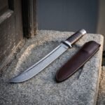Hand forged norse bowie knife on a concrete ledge, damascus clip point blade with a long fuller, stacked leather washer handle and a brown leather belt sheath
