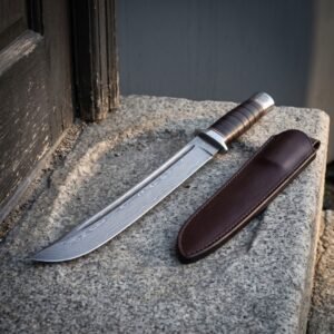 Hand forged norse bowie knife on a concrete ledge, damascus clip point blade with a long fuller, stacked leather washer handle and a brown leather belt sheath