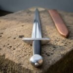 Close view of Norse dagger crossguard, black grip and round pommel on a stone block, double edged blade pointing away