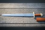 Hand forged norse sword with a broad fullered blade etched with twin ravens, brass brazil nut pommel and tan leather grip, laid on weathered grey decking with its scabbard.
