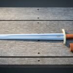 Hand forged norse sword with a broad fullered blade etched with twin ravens, brass brazil nut pommel and tan leather grip, laid on weathered grey decking with its scabbard.