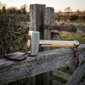 Hand forged norse war hammer with polished steel head cut with Elder Futhark runes and a pale ash haft, resting on a weathered farm gate with a leather head cover