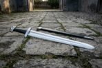 Hand forged norse warrior sword with a broad fullered polished blade, black leather grip and lobed steel pommel lying on a cobbled street beside its black scabbard