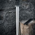 Hand forged pattern welded viking sword with a trilobate pommel and rune cut guard standing point down against a dark stone wall beside its black scabbard
