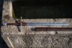 Hand forged ring pommel sword laid across a weathered stone trough, mirror polished blade, brown leather grip and wrapped ricasso, scabbard with two belt straps