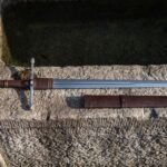 Hand forged ring pommel sword laid across a weathered stone trough, mirror polished blade, brown leather grip and wrapped ricasso, scabbard with two belt straps