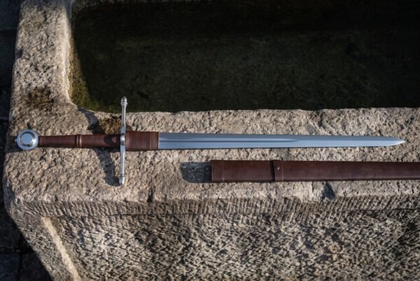 Hand forged ring pommel sword laid across a weathered stone trough, mirror polished blade, brown leather grip and wrapped ricasso, scabbard with two belt straps