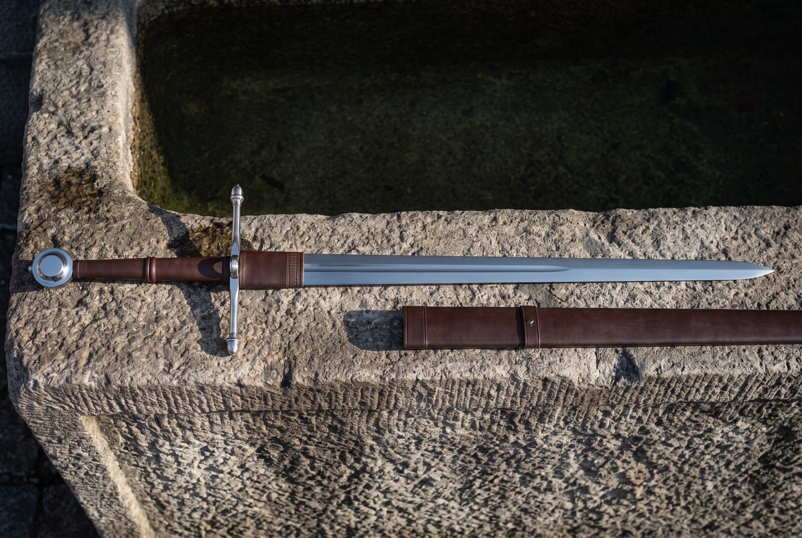 Hand forged ring pommel sword laid across a weathered stone trough, mirror polished blade, brown leather grip and wrappe Hand forged ring pommel sword laid across a weathered stone trough, mirror polished blade, brown leather grip and wrapped ricasso, scabbard with two belt straps