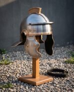 Hand forged roman legionary helmet in satin steel with bronze tone trim, engraved meander cheek guards and crest holder, shown on a wooden display stand outdoors