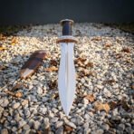 Hand forged Roman pugio dagger with raised midrib blade standing upright on gravel with fallen leaves and its sheath behind
