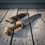 Hand forged Roman pugio dagger with solid brass guard and ribbed hardwood grip on wooden decking beside its brass fitted sheath