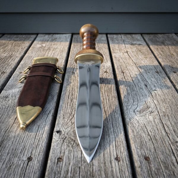 Hand forged roman pugio dagger with twin fullered waisted blade, brass guard and ribbed hardwood grip on grey decking beside a brass mounted leather sheath