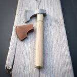 Hand forged rune axe lying on weathered grey planks, bearded high carbon head with an Elder Futhark band, pale ash haft, brass collar and a leather edge cover