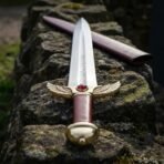 Hand forged rune engraved dagger with brass winged guard and red cabochon, viewed from the pommel on a mossy stone wall
