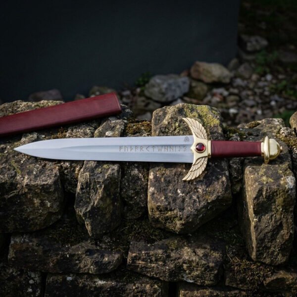 Hand forged rune engraved dagger with brass winged guard and red cabochon, full length side view on a stone wall with scabbard