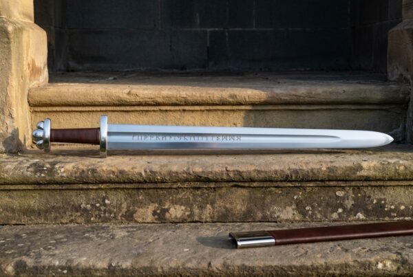 Hand forged rune engraved sword with the full Elder Futhark row cut along a satin polished blade lying on a sunlit stone step beside a brown leather scabbard