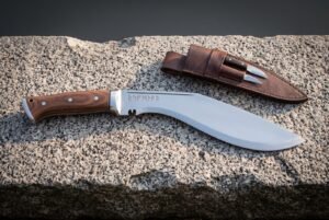 Hand forged rune kukri on a granite ledge, Elder Futhark line cut along the polished spine, dark rosewood scales, steel bolster and a leather sheath with companion knives