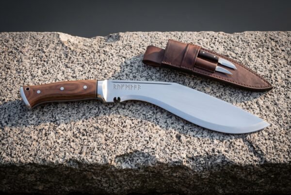 Hand forged rune kukri on a granite ledge, Elder Futhark line cut along the polished spine, dark rosewood scales, steel bolster and a leather sheath with companion knives