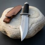 Sawback Bowie knife pointing down across a rounded stone, cord wrapped handle and brown leather sheath in view