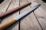 Hand forged seax with olive wood handle angled across old tabletop boards, close view of the grip with sheath beside