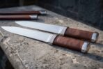 Hand forged seax pair with brown leather handles and steel fittings side by side on stone, fitted sheaths above