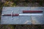 Hand forged side ring sword on a grey slate slab, polished steel ring guard and burgundy leather grip stamped with a raven, matching leather scabbard alongside