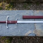 Hand forged side ring sword on a grey slate slab, polished steel ring guard and burgundy leather grip stamped with a raven, matching leather scabbard alongside