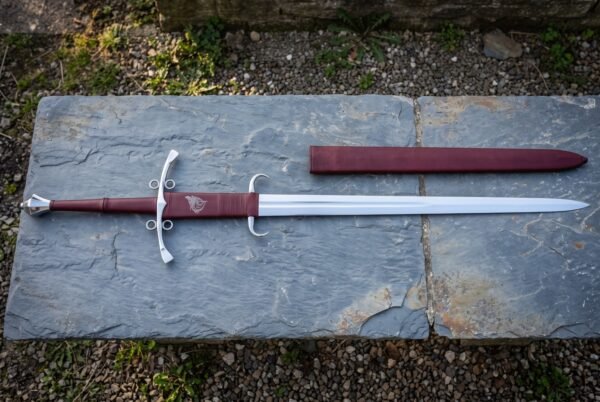 Hand forged side ring sword on a grey slate slab, polished steel ring guard and burgundy leather grip stamped with a raven, matching leather scabbard alongside