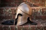 Hand forged steel Corinthian Greek helmet with bronze tone finish and hammered crown, side angle view resting on red brick steps