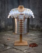 Hand forged steel lorica segmentata cuirass with articulated shoulder guards and brass buckles, displayed on a wooden stand against a weathered stone wall in daylight.