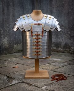 Hand forged steel lorica segmentata cuirass with articulated shoulder guards and brass buckles, displayed on a wooden stand against a weathered stone wall in daylight.