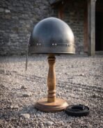 Hand forged steel medieval nasal helmet with rounded skull and riveted browband on a wooden stand in a gravel yard, side view
