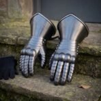Hand forged steel plate gauntlets with articulated finger lames standing on stone steps beside a pair of black gloves