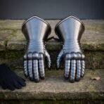 Hand forged steel plate gauntlets with engraved cuffs seen face on, fingers down on mossy stone steps with black gloves