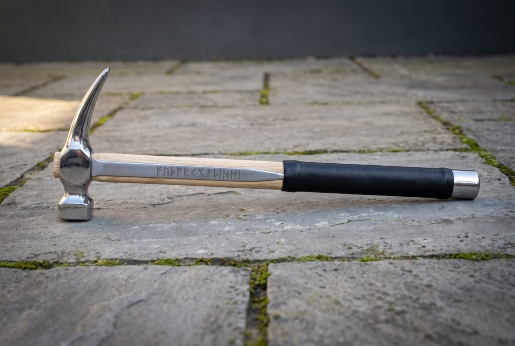 Hand forged steel rip claw hammer with a polished head, hickory handle carrying a cut rune line, black rubber over grip and a steel end cap on stone paving.