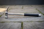 Hand forged steel rip claw hammer with a polished head, hickory handle carrying a cut rune line, black rubber over grip and a steel end cap on stone paving.