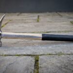 Hand forged steel rip claw hammer with a polished head, hickory handle carrying a cut rune line, black rubber over grip and a steel end cap on stone paving.