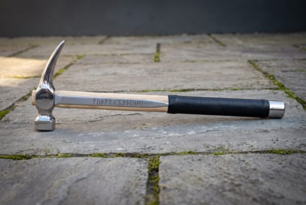 Hand forged steel rip claw hammer with a polished head, hickory handle carrying a cut rune line, black rubber over grip and a steel end cap on stone paving.