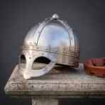 Hand forged steel Viking spectacle helmet with riveted skull and brass browband, angled view on a stone bench by a leather liner