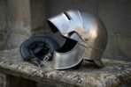 Hand forged steel visored medieval helmet with perforated bevor and raised visor on a stone bench beside its padded leather liner