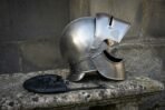 Hand forged steel visored medieval helmet in side profile with the visor lifted, resting on stone next to its black leather liner
