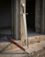 Hand forged sword with tan leather grip and engraved disc pommel leaning in a stone doorway with a russet leather scabbard