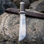 Hand forged tanto knife with stacked leather handle and steel guard, close view of the fullered blade on a boulder with its sheath