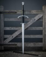 Hand forged two handed longsword with parrying hooks and a ring terminal crossguard standing against a weathered farm gate above its black leather scabbard on gravel