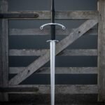 Hand forged two handed longsword with parrying hooks and a ring terminal crossguard standing against a weathered farm gate above its black leather scabbard on gravel