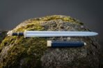 Hand forged viking arming sword with a fullered polished blade, brass knotwork guard, lobed pommel and blue leather grip lying on a mossy boulder with its scabbard