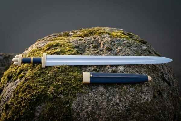 Hand forged viking arming sword with a fullered polished blade, brass knotwork guard, lobed pommel and blue leather grip lying on a mossy boulder with its scabbard