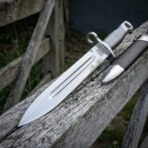 Hand forged Viking bayonet with ribbed steel grip and etched fuller on a weathered fence rail beside its black scabbard
