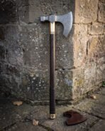 Hand forged Viking bearded axe with etched knotwork head, engraved brass collar and leather wrapped haft leaning against a lichen covered sandstone wall