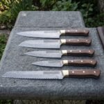 Hand forged Viking chef knife set of five Damascus blades with knotwork engraved panels, walnut handles and brass bolsters, laid on slate beside a brown leather knife roll.